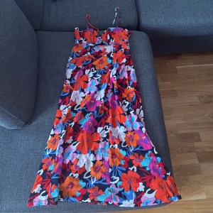Färgglad blommig långklänning BikBok - Supersnygg långklänning från BikBok med tunna axelband och färgstarkt blommigt mönster i rött, lila, blått, orange och vitt. Klänningen har figurnära överdel och v-ringning, perfekt för sommarens alla tillfällen.