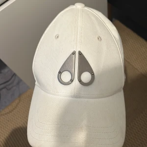 Vit Moose Knuckels keps - Vit keps i bomull från 1017 ALYX 9SM med deras ikoniska metallogo framtill. Justerbar rem baktill med metallspänne. Snygg och minimalistisk design som sticker ut med den unika logodetaljen.