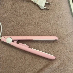 Rosa plattång för håret - Söt plattång i ljusrosa med vita detaljer och sladd med EU-kontakt. Kompakt och smidig design som passar perfekt för styling av håret. Plattorna är vita och själva tången är gjord av plast.