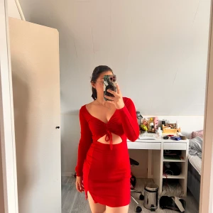 Röd kort klänning med cut-out och snörning - Säljer en röd kort klänning med lång ärm, snygg cut-out vid magen och snörning framtill. Klänningen har v-ringning, liten slits på sidan och figurnära passform. Perfekt för dig som vill sticka ut med en trendig look.