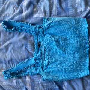 Turkos smockad linnetopp - Supersöt turkos linnetopp med smockad struktur och breda axelband. Toppen har volangkant upptill och nertill för en extra gullig touch. Perfekt för varma dagar och enkel att styla med jeans eller kjol.