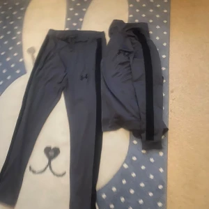 Grå träningssett från Under Armour - Säljer ett par grå träningsbyxor från Under Armour med svarta sidopaneler och dragsko i midjan. Byxorna är i mjukt och stretchigt syntetmaterial, perfekta för träning eller chill. Klassisk logga på låret och smal passform. Använd kanske 2-3 veckor inte mer.