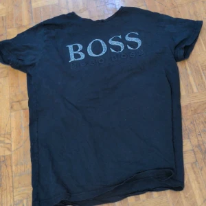 Svart t-shirt från Hugo Boss - Svart t-shirt från Hugo Boss med stort BOSS-tryck på bröstet där bokstäverna har ett mönster med små logotyper. Klassisk passform och rund hals. Perfekt för dig som gillar stilrena och snygga märkesplagg. Vet inte om den är äkta eller inte fick den som en prisent