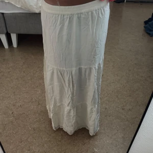 Vit långkjol med svarta fina sömmar längst ner på kjolen - Cremevit långkjol med volangdetaljer och elastisk midja. Kjolen har flera sömmar och svarta dekorativa ränder längst ner. Lätt och flowy känsla, perfekt för varma dagar. Enkel att matcha med olika toppar för en chill och stilren outfit. Tyvärr bara använt en gång, når till marken för mig som är ca 165 med långa ben. 