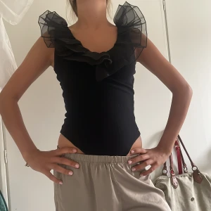 Svart body med volangkrage - Supersnygg svart body med transparent volangkrage i mesh som ger en dramatisk look. Ärmlös modell med djup urringning och figurnära passform. Perfekt för dig som vill sticka ut med en unik topp.