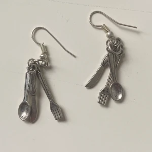 Örhängen med bestick i silverfärg - Unika örhängen med små hängande bestick – kniv, gaffel och sked – i silverfärgad metall. Perfekt för dig som gillar quirky accessoarer och vill sticka ut. Lätta att bära och ca 5 cm långa. Säljs som par.