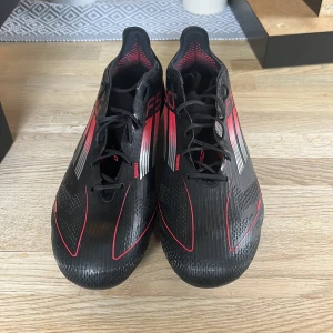 Adidas F50 Elite FG fotbollsskor - Säljer ett par Adidas F50 Elite FG fotbollsskor i svart med röda och silvriga detaljer. Skorna är bara provade, säljs pågrund av fel storlek. Allt är med, skor, skopåsen och extra sulor. Nypris 2300kr