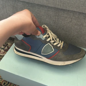 Philippe Model blå/grå sneakers strl 42 - Snygga sneakers från Philippe Model i blått och grått med röda detaljer. Ovandel i mocka och textil, vit och svart sula samt vita snören. Skorna har en sportig vibe med rund tå och coola färgblock. Perfekta för dig som gillar streetstyle och vill sticka ut.