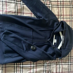 Marinblå vindjacka från C.P. Company - Snygg marinblå vindjacka från C.P. Company med huva, dragkedja och klassisk linsdetalj på ärmen. Jackan har praktiska fickor och en clean look som passar perfekt till streetwear. Materialet är mjukt och lätt, perfekt för vår och höst.