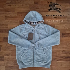 Burberry Hoodie - Letar du efter en stilren och lyxig hoodie? Denna helt nya grå Burberry-hoodie kombinerar elegans med hög komfort. Den har Burberrys ikoniska rutmönster i fodret och en diskret logga på bröstet för en exklusiv touch. Tillverkad av mjukt, slitstarkt material och utrustad med dragkedja samt huva – både praktisk och stilfull.  📌 Skick: Ny med taggar. 📦 Snabb leverans – skickas samma dag! .