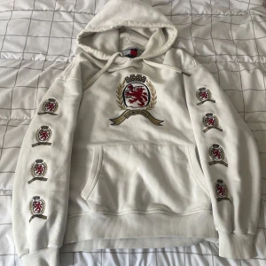 Vit hoodie Tommy Jeans med brodyr - Snygg vit hoodie från Tommy Jeans med broderat lejonemblem på bröstet och flera broderade märken längs båda ärmarna. Klassisk huva med snörning och stor känguruficka framtill. Perfekt för dig som gillar streetwear med preppy touch.