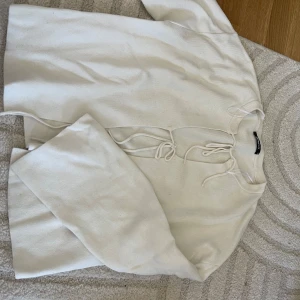 Vit stickad tröja med snörning - Vit stickad tröja med snygg snörning framtill vid halsen. Tröjan har lång ärm och en avslappnad passform, perfekt för lager på lager. Ribbad kant vid halsen ger en extra detalj. Enkel att matcha med jeans eller kjol.