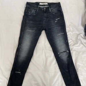 Svarta Replay jeans med slitningar - Snygga svarta jeans från Replay med slitna detaljer och lätt tvättad look. Modellen har klassisk femficksdesign, smal passform och coola slitningar på benen. Perfekta för dig som gillar en edgy stil och vill ha jeans med attityd.