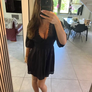 Svart kort klänning med spetsdetalj - Supersöt svart kort klänning med v-ringning och fina spetsdetaljer längs urringningen. Ärmarna och midjan är tajtare runt om. Tunt, lätt tyg som känns luftigt och skönt.