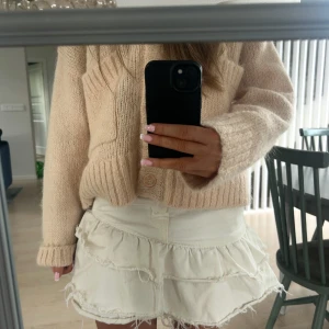 Vit volangkjol i jeans med råa kanter - Supersöt vit kortkjol i jeans med volanger och råa kanter för en trendig look. Kjolen har klassisk jeansmidja med knapp och dragkedja, samt flera lager volanger som ger extra volym och edge.