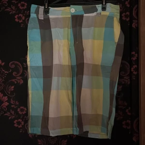 Rutig shorts från Cover Streetwear - Färgglada rutiga shorts från Cover Streetwear i blått, gult, grönt, grått och vitt. Klassisk passform med knapp och dragkedja framtill samt sidfickor. Perfekta för sommardagar när du vill sticka ut lite extra.