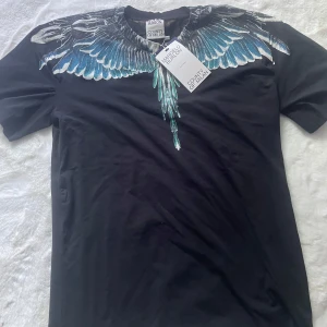 Svart t-shirt Marcelo Burlon med vingar - Svart t-shirt från Marcelo Burlon County of Milan med ikoniskt vingtryck i blått och vitt över axlar och bröst. Klassisk rund hals och korta ärmar. Grym statement-piece för dig som gillar streetwear och unika prints.