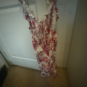 Blommig långklänning med volang - Romantisk långklänning med blommigt mönster i rött och rosa på vit botten. Klänningen har volangdetaljer längs urringningen och axelbanden samt knyt framtill. Perfekt för sommarens alla tillfällen.