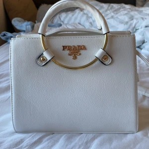 Look alike väska från Pr@da - Snygg vit handväska som är köpt på en marknad i Spanien för några år sedan. Väskan är i skinn och har flera fack med dragkedja inuti, perfekt för att hålla ordning på dina grejer. Klassisk boxig form som ger en lyxig vibe. Axelremsband medföljer!