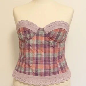 Rutig korsettopp med spetsdetaljer - Superfin korsettopp i rutigt mönster med lila, rosa och gröna toner. Toppen har spetskant upptill och nedtill som ger en söt touch. Axelbandslös design och figurnära passform, perfekt för att sticka ut lite extra.