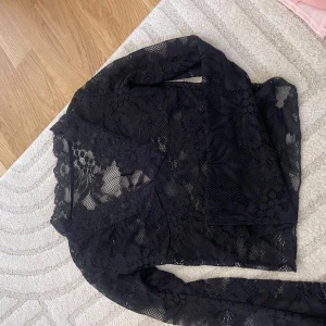 Svart spetsblus med lång ärm - Säljer en svart blus i genombruten spets med blommigt mönster. Toppen har långa ärmar och v-ringad halsringning med fina spetsdetaljer. Perfekt för dig som gillar en elegant och trendig look.