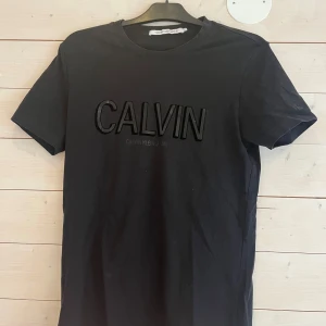 Svart t-shirt från Calvin Klein Jeans - Svart t-shirt från Calvin Klein Jeans med stor, svart CALVIN-logga framtill. Klassisk rund halsringning och korta ärmar. Tillverkad i mjuk bomull för en skön känsla och clean look. Perfekt för dig som gillar stilrena och trendiga plagg.