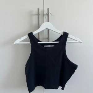 Svart croppad topp - En svart croppad topp från 157 med breda axelband och rund halsringning. Toppen är ribbad och har en enkel, stilren design som passar perfekt till jeans eller kjol. Materialet är mjukt och stretchigt för en bekväm känsla.