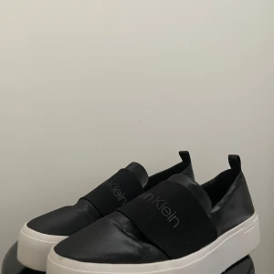 Svarta sneakers Calvin Klein - Stilrena svarta slip-on sneakers från Calvin Klein med bred resår över foten och logga framtill. Skorna har vit platt sula och ovandel i syntetmaterial med diskret glans. Perfekta för en modern look.