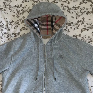 Burberry zip - Snygg grå hoodie från Burberry med klassiskt rutigt foder i huvan. Tröjan har hel dragkedja, snörning i huvan och broderad logga på bröstet. Tillverkad i mjuk bomullsmix, perfekt för chill dagar. Stilsäker detalj med Burberrys ikoniska mönster i huvan.