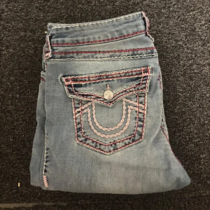Ljusblå bootcut jeans med rosa sömmar - Säljer ett par ljusblå jeans från true religion med bootcut passform. Jeansen har unika rosa och vita kontrastsömmar, broderade detaljer på bakfickan och klassisk femficksdesign. Materialet är jeans och de har låg midja. Perfekta för dig som vill sticka ut lite extra. Nypris ca 2500kr. Gått sönder lite längst ner där men som inte syns eller som man tänker på 