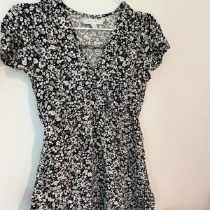 Svartvit blommig klänning Amisu - Svartvit kortklänning från Amisu med blommigt mönster, v-ringning med överlappning och korta flowiga ärmar. Klänningen har en lätt figursydd passform som sedan går ut lite i midjan. Tunt och skönt material som känns luftigt. Säljer då den är alldeles för kort på mig.