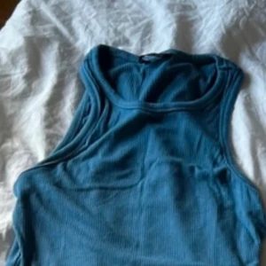 Blå ribbad linnetopp - Säljer en blå ribbad linnetopp med rund halsringning och ärmlös design. Toppen är croppad och har en enkel, stilren look som passar perfekt till jeans eller kjol. Materialet är mjukt och stretchigt för en bekväm passform.