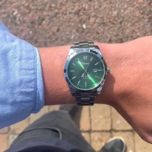 Grön klocka från Olevs med stållänk - Stilren klocka från Olevs med grön urtavla och silverfärgad länk i metall. Klockan har tydliga index, datumvisning och klassisk rund form. Perfekt accessoar för dig som vill sticka ut med en snygg detalj på handleden.