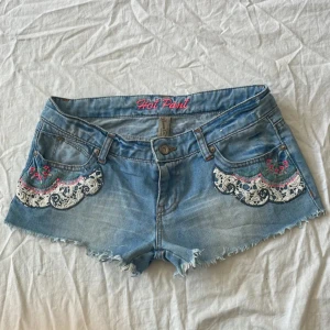 Lågmidjade Jeansshorts - Snygga ljusblå jeansshorts från Hot Pant med råa kanter och broderade detaljer i vitt, rosa och blått på fickorna framtill. Shortsen har låg midja och klassisk femficksdesign. Perfekta för sommarens alla äventyr!