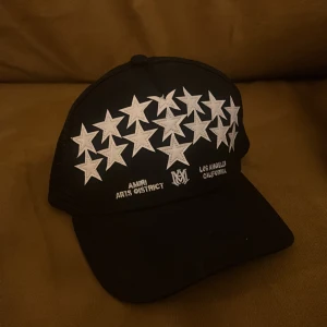 Svart keps från Amiri med stjärnor - Svart keps från Amiri med vit broderad stjärndesign framtill och texten 'AMIRI ARTS DISTRICT' samt 'LOS ANGELES CALIFORNIA'. Kepsen har mesh på sidorna och justerbar snapback baktill. Snygg streetwear-vibe och tydlig Amiri-logga invändigt.