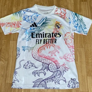 Real Madrid x Adidas draktröja - Säljer en unik Real Madrid fotbollströja från Adidas med färgglad drakdesign och molnmönster över hela tröjan. Vit bas med detaljer i blått, rött, gult och grönt. Klassisk rund hals och korta ärmar. Materialet är lätt och andas, perfekt för träning eller match.