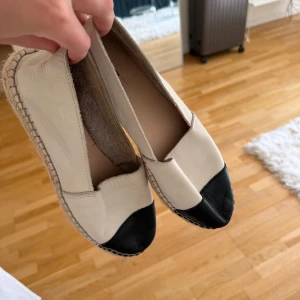 Beige och svarta espadrillos i skinn - Snygga espadrillos i beige skinn med svart tåparti och klassisk flätad sula. Skorna har en rund tå och platt sula, perfekta för en chill och stilren look. Lätta att matcha till jeans eller klänning för en avslappnad vibe.