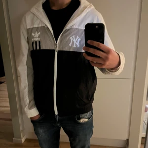 Adidas x NY Yankees vindjacka svart/vit - Snygg vindjacka från Adidas i samarbete med New York Yankees. Jackan är svart, grå och vit med huva, dragkedja och loggor på bröstet och ryggen. Lätt material i polyester, perfekt för vår och höst. Klassisk streetstyle med raka linjer och sportig vibe. Ena fickans dragkedja funkar inte.
