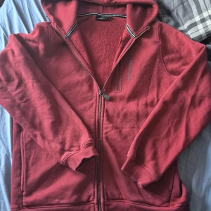 Röd hoodie från Sail Racing med dragkedja - Säljer en röd hoodie från Sail Racing med hel dragkedja och huva. Tröjan har broderad logga på bröstet, två fickor fram och är fodrad med mjuk fleece på insidan. Materialet är en mix av bomull och polyester. Perfekt för kyliga dagar.