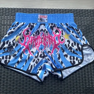 Blå leopardmönstrade Muay Thai-shorts - Snygga Muay Thai-shorts från US Angels med blå och svarta leopard- och zebramönster, bred resår i midjan och coola detaljer som stjärnor och rosa tryck framtill. Perfekta för dig som vill sticka ut på träningen.