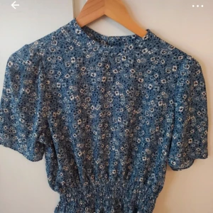 Blommig blå klänning Gina Tricot - Supersöt blå blus från Gina Tricot med små vita och svarta blommor. Klänningen har korta ärmar, rund hals och resår i midjan för en snygg passform. Perfekt för sommarens alla tillfällen!