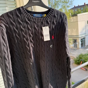 Mörkgrå kabelstickad tröja Polo Ralph Lauren - Kabelstickad mörkgrå tröja från Polo Ralph Lauren med klassisk rund hals och röd broderad logga på bröstet. Tröjan är långärmad och tillverkad i mjuk bomull, perfekt för lager-på-lager och snygg till jeans. Priset kan diskuteras vid snabb affär