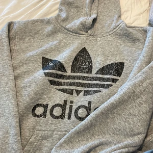Grå Adidas hoodie med stor logga - Säljer en grå hoodie från Adidas med stor svart Trefoil-logga och text framtill. Tröjan har en luva och en ficka framtill. Tröjan är dock sönder på ärmmudden men inget som syns. Tröjan är från Grekland så ej äkta