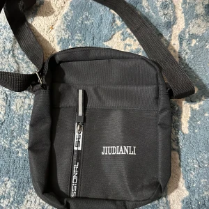 Svart axelväska från Jiudianli - En stilren svart axelväska från Jiudianli med justerbar rem och dragkedja framtill. Väskan är tillverkad i slitstark polyester och har ett enkelt, clean look utan mönster. Perfekt för att bära dina viktigaste grejer när du är på språng.