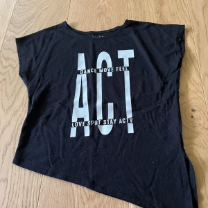 Svart topp med tryck ACT - Svart kortärmad topp med stort vitt tryck framtill där det står 'ACT' och texten 'DANCE MOVE FEEL' samt 'LOVE SPRT STAY ACTV'. T-shirten har en loose passform och är tillverkad i mjuk bomull, perfekt för en sportig och avslappnad look.