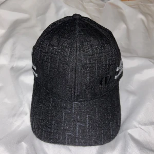Svart keps från Christian Dior - Snygg svart keps från Christian Dior med diskret mönster i tyget och broderad CD-logga på sidan. Kepsen har en grå och svart detalj med texten 'Christian Dior Paris' på sidan. Tillverkad i ett strukturerat tyg med exklusiv känsla.