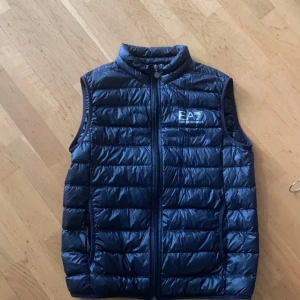 Blå dunväst från Emporio Armani EA7 - Snygg svart dunväst från Emporio Armani EA7 med quiltad design och hög krage. Västen har dragkedja framtill och logga på bröstet. Perfekt att slänga över en hoodie eller tröja för extra värme. Klassisk och stilren look.