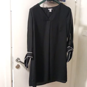 Svart klänning med vita detaljer H&M - Stilren svart kortklänning från H&M med v-ringning och långa ärmar. Ärmarna har snygga vita kantband och breda manschetter som ger en cool touch. Klänningen har rak passform och är tillverkad i ett lätt, mjukt tyg som faller snyggt.