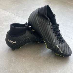 Nike Air Zoom svarta fotbollsskor - Svarta Nike Air Zoom fotbollsskor med högt skaft och snörning. Skorna har en sportig design med subtila silverdetaljer och gula dobbar undertill. Ovandelen är i syntetmaterial med diskret mönster och Nike-logga på sidan. Perfekta för dig som vill ha stil på planen.