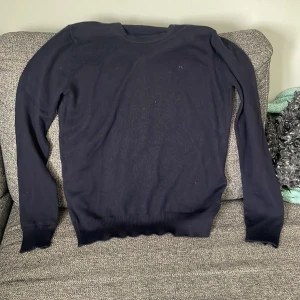 Mörkblå stickad tröja från J Lindeberg  - Klassisk mörkblå stickad tröja från JL med diskret logga på bröstet. Tröjan har rund halsringning, långa ärmar och ribbade muddar vid ärmslut och nederkant. Perfekt för en stilren och enkel look.